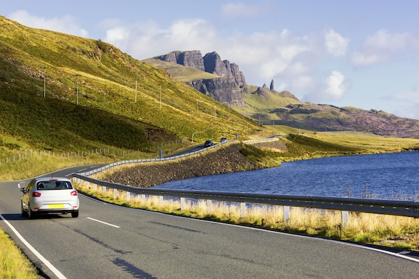 a silver car driving past hills next to an expanse of water in the UK